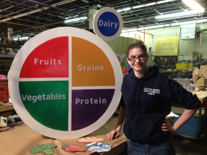 Ms. O'Brien stands beside an oversized rolling MyPlate prop constructed for a touring education production, Nick Nutrition.